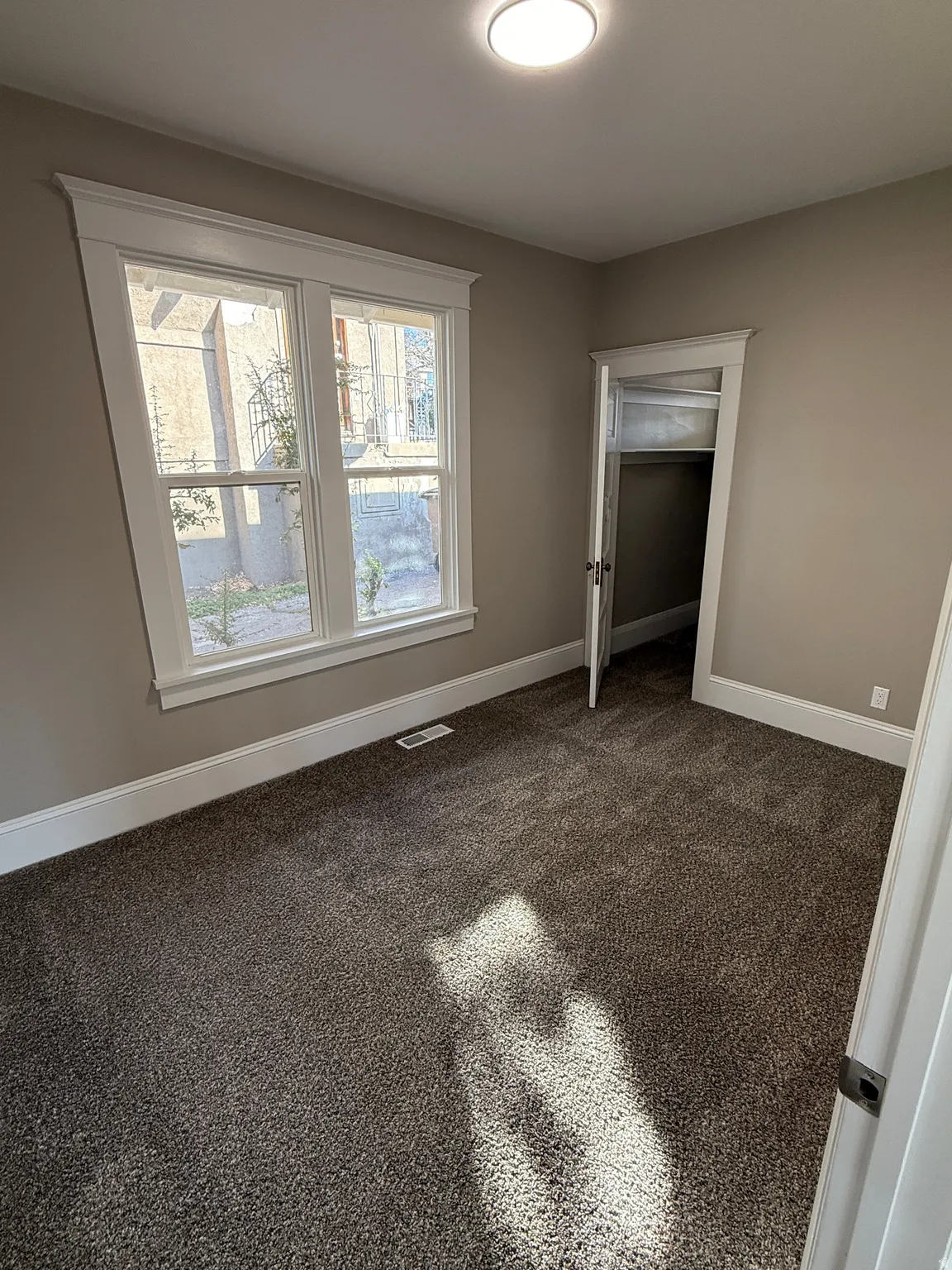 Wall-to-wall carpeting with a neutral finish, featuring a double-hung window with white trim, a built-in closet with a bi-fold door, and a flush-mount ceiling light fixture