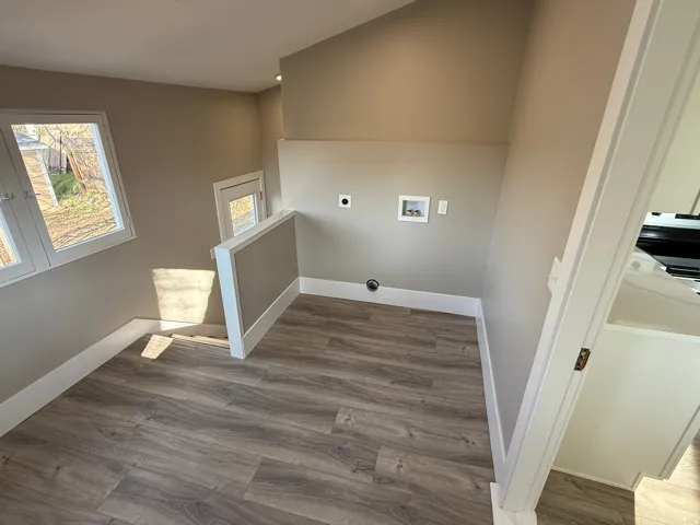 Dedicated utility space featuring wood-finish flooring, multiple windows, a half-wall partition, electrical outlets, and plumbing hookups