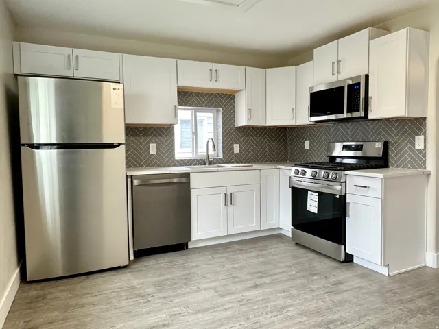 Modern kitchen featuring white shaker cabinetry, stainless steel appliances, herringbone tile backsplash, light countertops, and wood-finish flooring