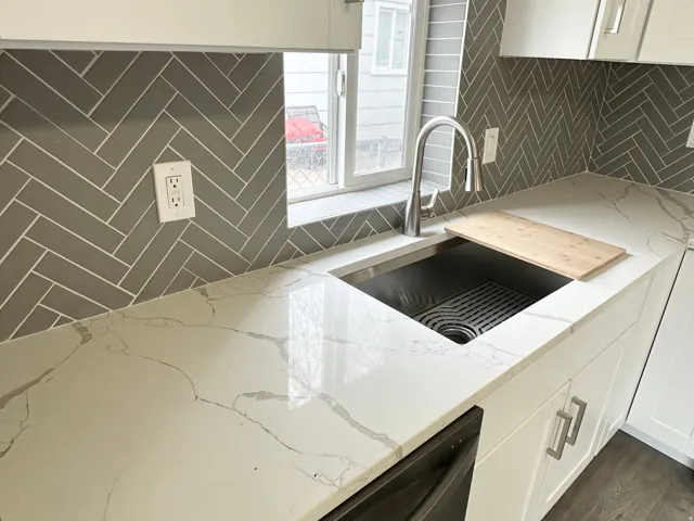Kitchen area featuring white cabinetry with silver-tone hardware, a stainless steel sink with a gooseneck faucet, and a light-toned countertop with veining