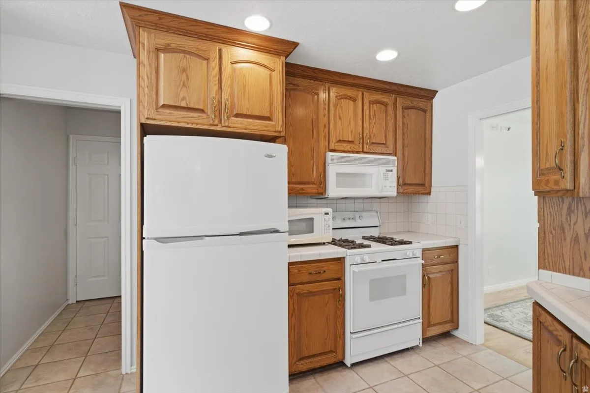 Kitchen featuring wood cabinetry, white appliances, tile countertops, tile backsplash, and recessed lighting