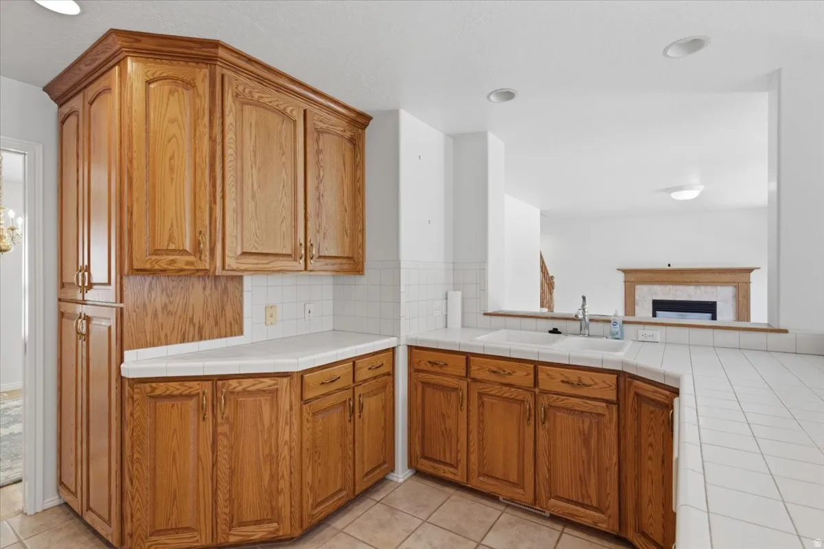Kitchen featuring extensive wood cabinetry, white tiled countertops, white tile backsplash, an integrated sink, and a pass-through window to an adjacent room with a fireplace