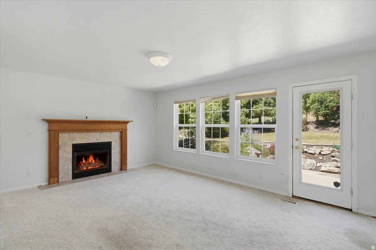Living space featuring a tiled fireplace with wood mantel surround, light-colored carpeting, multiple windows, and a glass-paneled exterior door