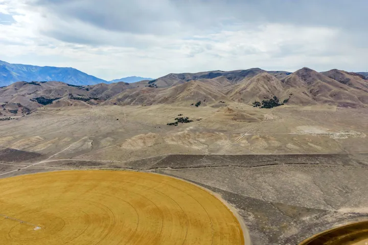 Expansive rural landscape featuring a prominent circular field, surrounding natural terrain, and distant mountains under a cloudy sky