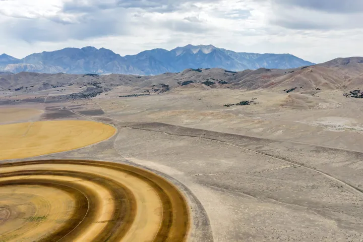 Expansive agricultural land featuring circular crop patterns, open fields, and distant mountain ranges under a cloudy sky