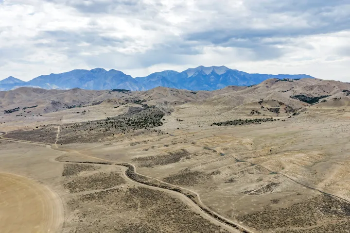 Expansive high desert landscape featuring a winding path, distant mountain range, and scattered shrubbery under a cloudy sky