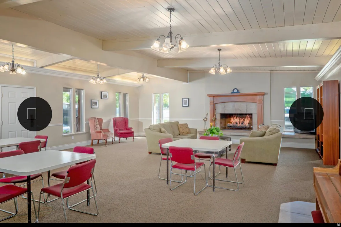 Spacious common area featuring a stone fireplace with a wood mantel, exposed ceiling beams, multiple windows, and a combination of ambient and task lighting