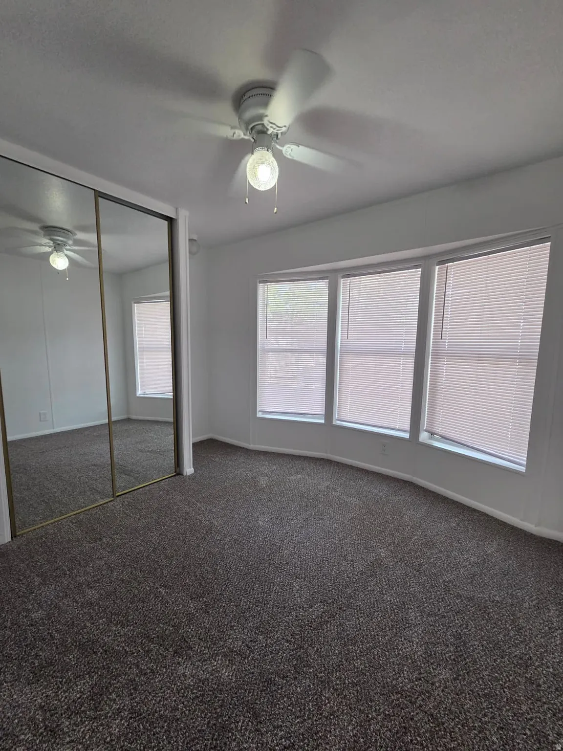 Bedroom featuring a neutral color palette, dark gray carpeting, a bay window with horizontal blinds, a ceiling fan with integrated lighting, and mirrored closet doors
