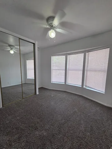 Bedroom featuring a neutral color palette, dark gray carpeting, a bay window with horizontal blinds, a ceiling fan with integrated lighting, and mirrored closet doors