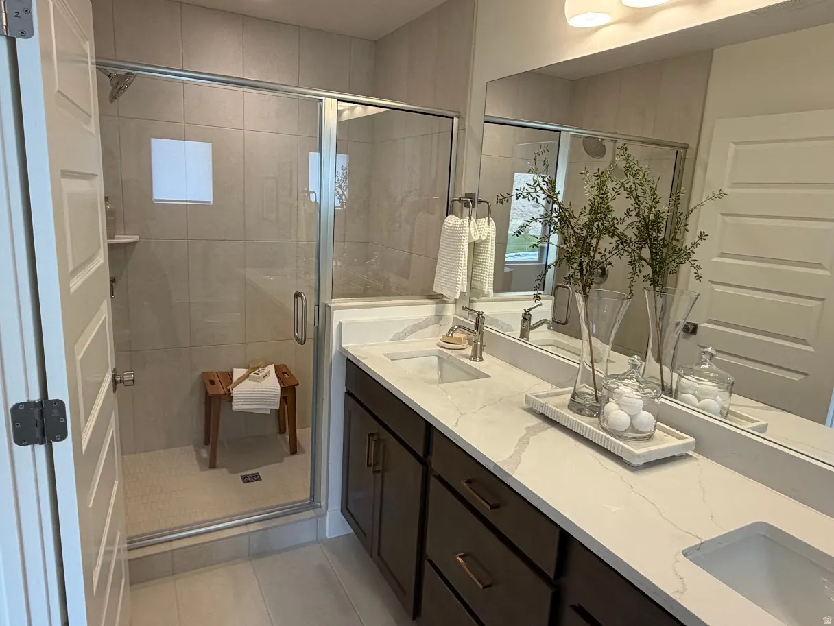 Bathroom featuring a dual vanity with undermount sinks, quartz countertops, dark wood-finish cabinetry, a large wall mirror, and a glass-enclosed shower with tiled walls and a built-in shower shelf