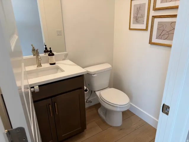 Powder room featuring a single vanity with a white countertop, built-in sink, and bronze-finish hardware, complemented by wood-finish flooring and a toilet