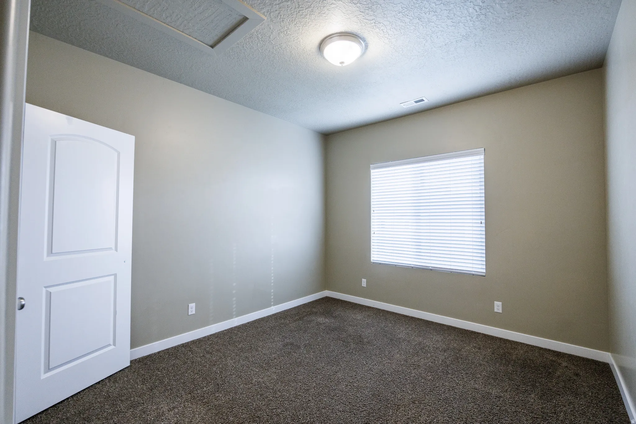 Neutral-toned room featuring a large window with horizontal blinds, textured ceiling, and a flush-mount light fixture