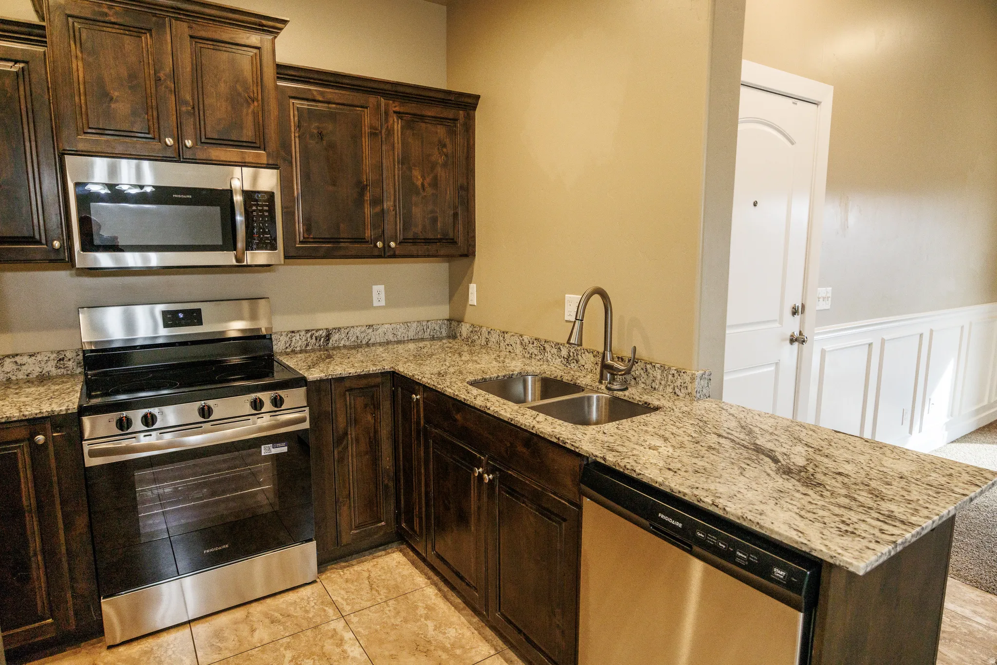 Kitchen featuring dark wood cabinetry, stainless steel appliances, speckled stone countertops, a double basin sink with a high arc faucet, and tile flooring