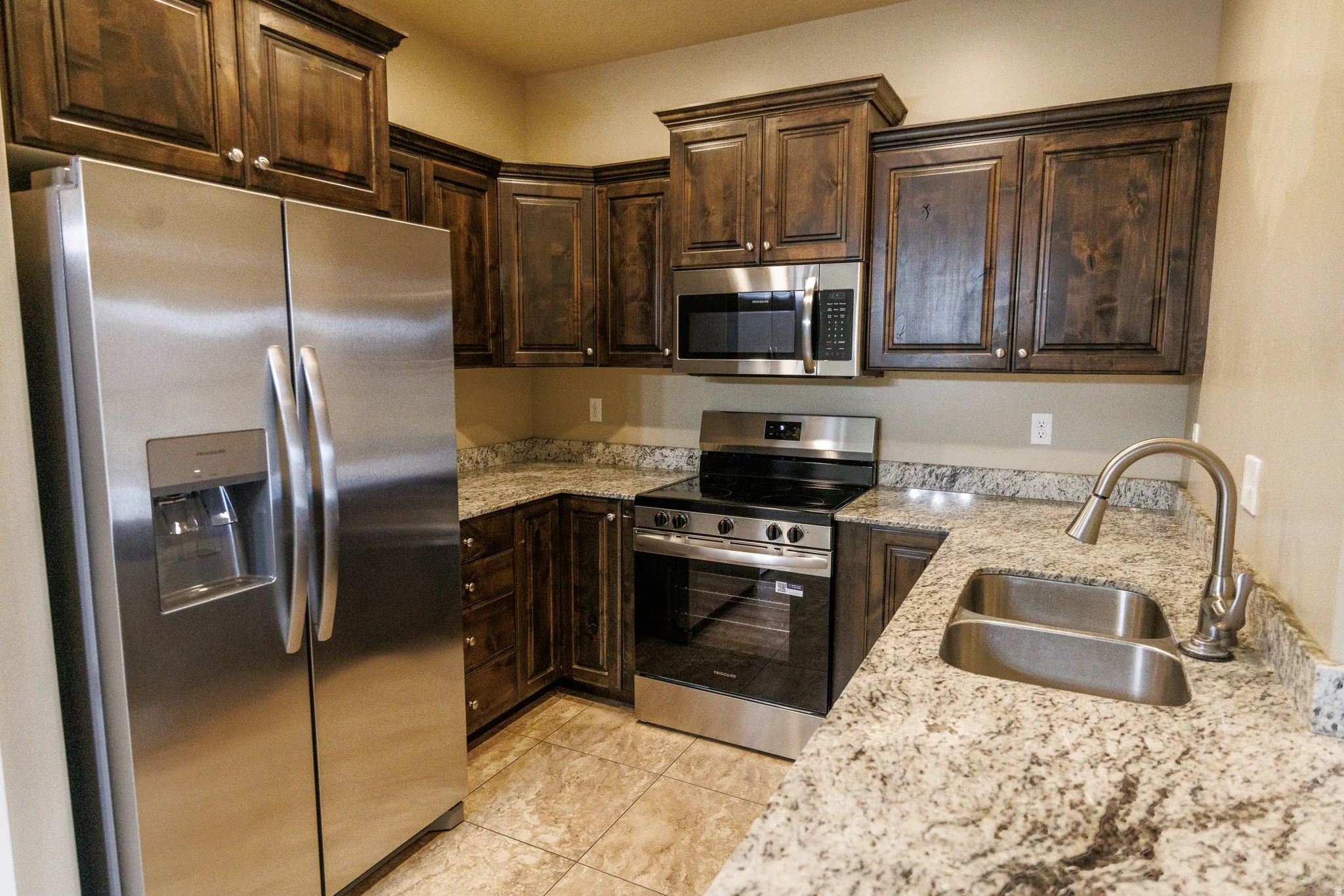 Kitchen featuring dark wood cabinetry, light-toned granite countertops, stainless steel appliances, a double basin sink with a brushed nickel faucet, and ceramic tile flooring