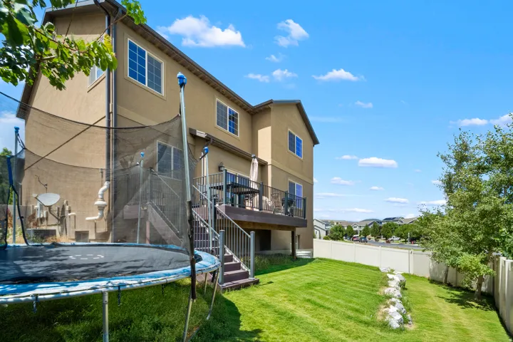 Rear exterior featuring a spacious elevated deck with dark railings, a lower-level concrete patio, a manicured lawn, and a light-colored privacy fence
