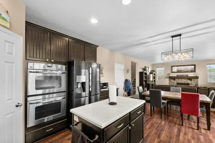 Open-concept living area featuring wood-finish flooring throughout, a kitchen island with a white countertop, dark wood cabinetry, and stainless steel double ovens