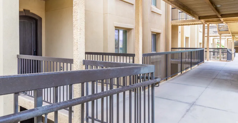 Covered exterior walkway featuring a smooth concrete surface, dark metal railings, and a stucco-textured building facade