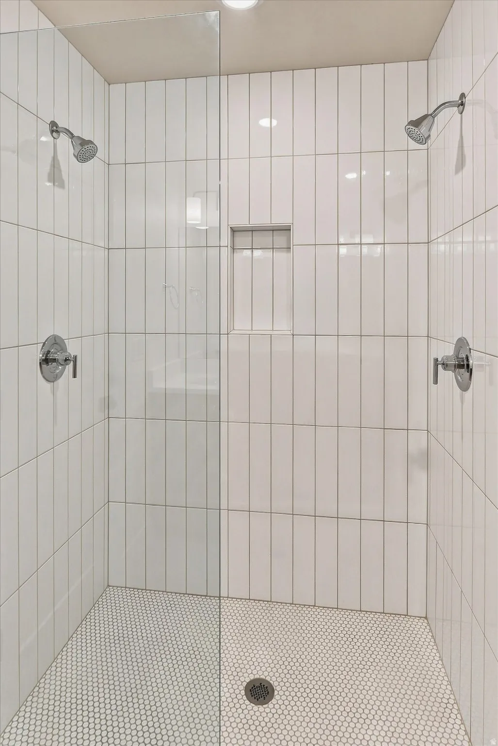 Shower enclosure featuring white vertical subway tile, dual shower heads, a built-in shower niche, and white hexagonal mosaic floor tiles