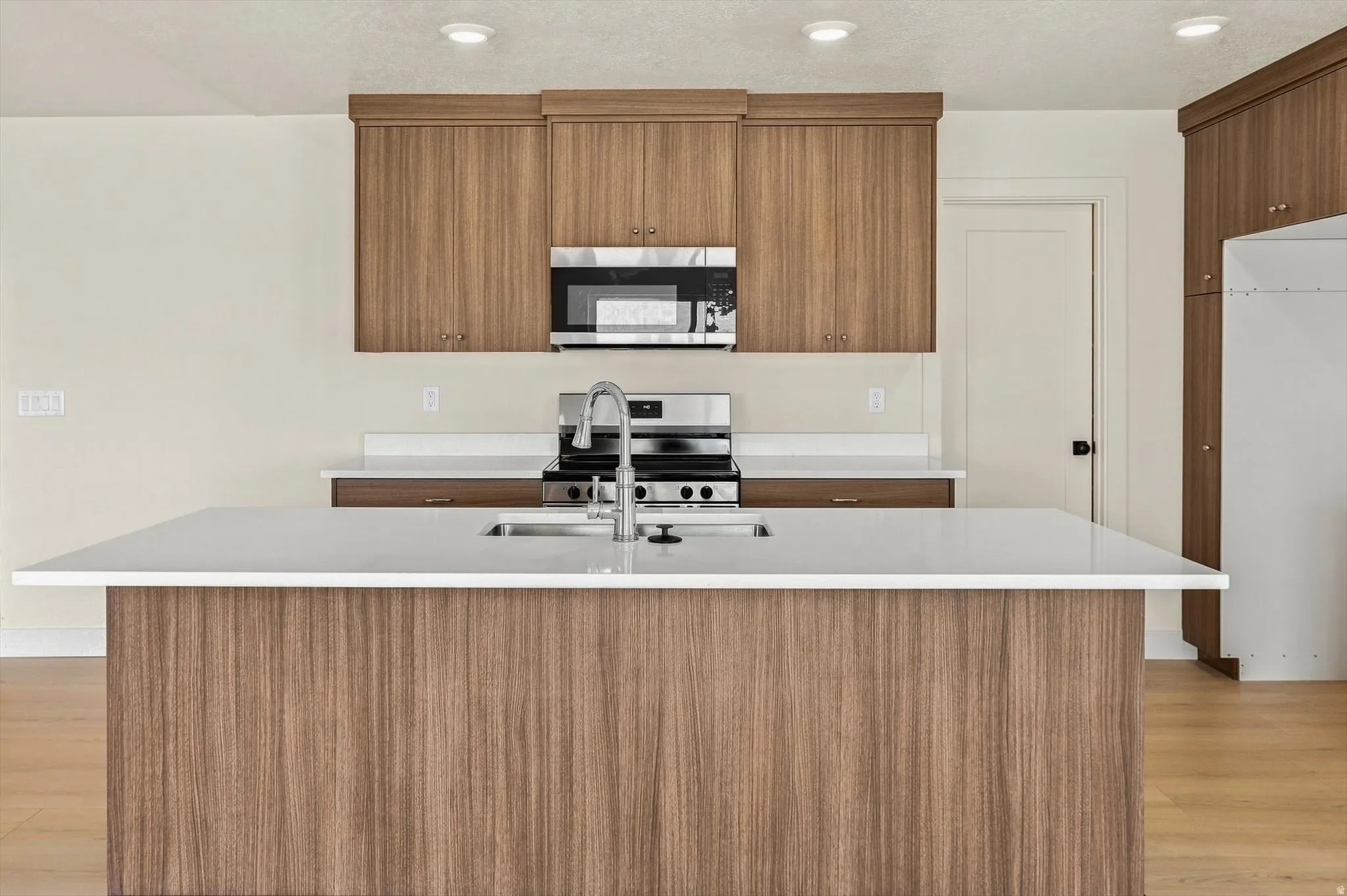 Kitchen featuring a large central island with an integrated sink, white countertops, wood-finish cabinetry, stainless steel appliances, and recessed lighting