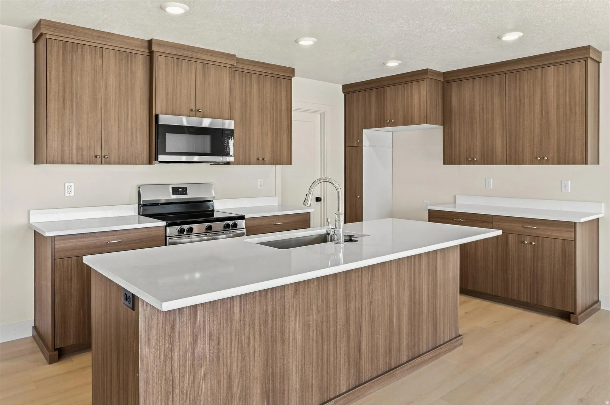 Modern kitchen featuring a central island with a built-in sink and faucet, wood-finish cabinetry, stainless steel appliances, light countertops, and recessed lighting