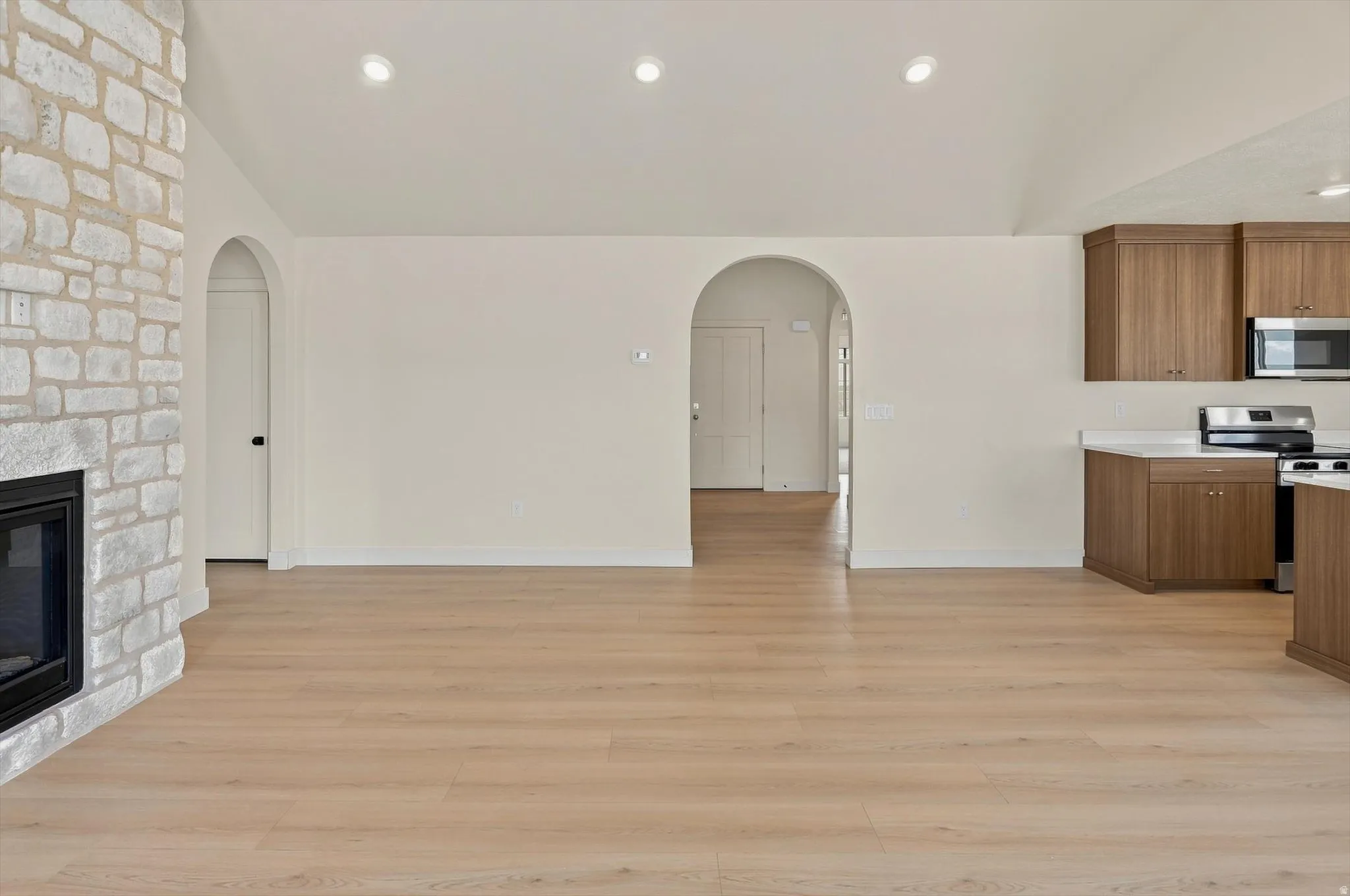 Spacious room featuring light wood-finish flooring, a stone-clad fireplace with a black firebox, multiple recessed lights, a vaulted ceiling, and arched doorways
