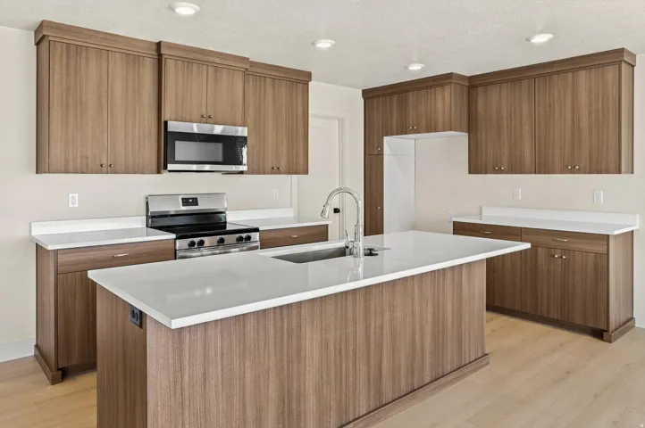 Modern kitchen featuring a central island with a built-in sink and faucet, wood-finish cabinetry, stainless steel appliances, light countertops, and recessed lighting