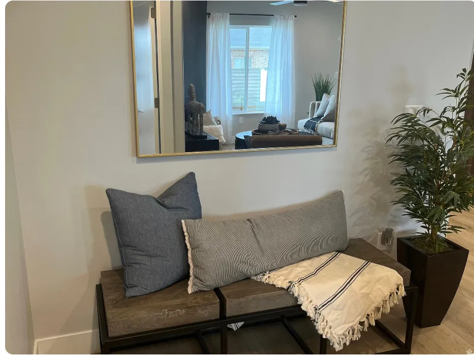 Wall-mounted gold-framed mirror reflecting a room with a window, light-toned walls, and wood-finish flooring