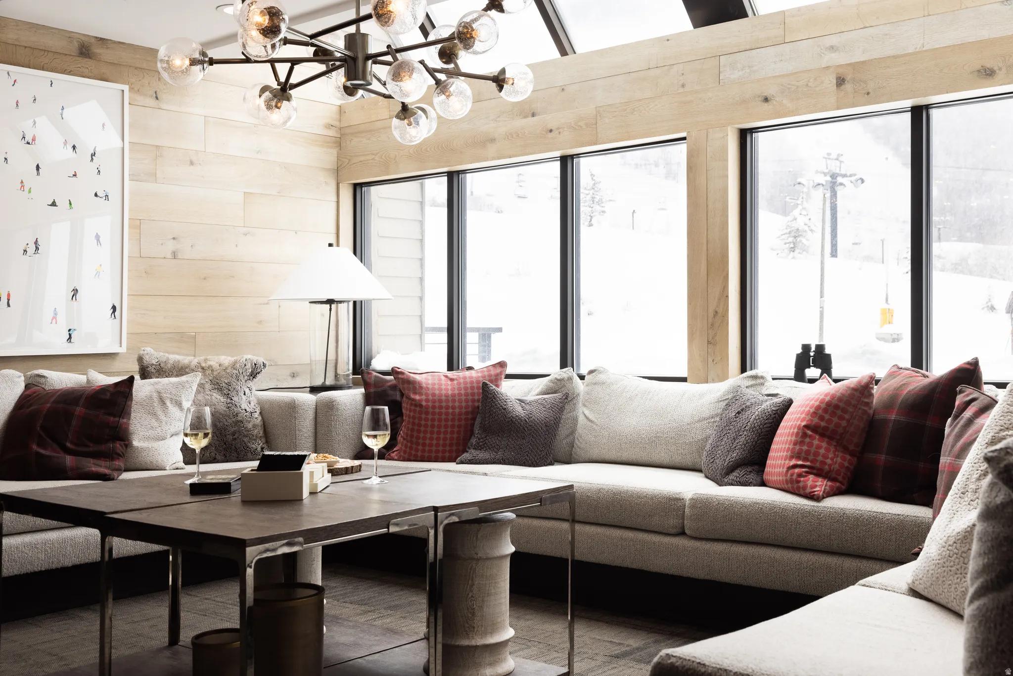 Open-concept living area featuring a vaulted ceiling with skylights, extensive wood-finish wall paneling, and large picture windows showcasing a snowy mountain landscape