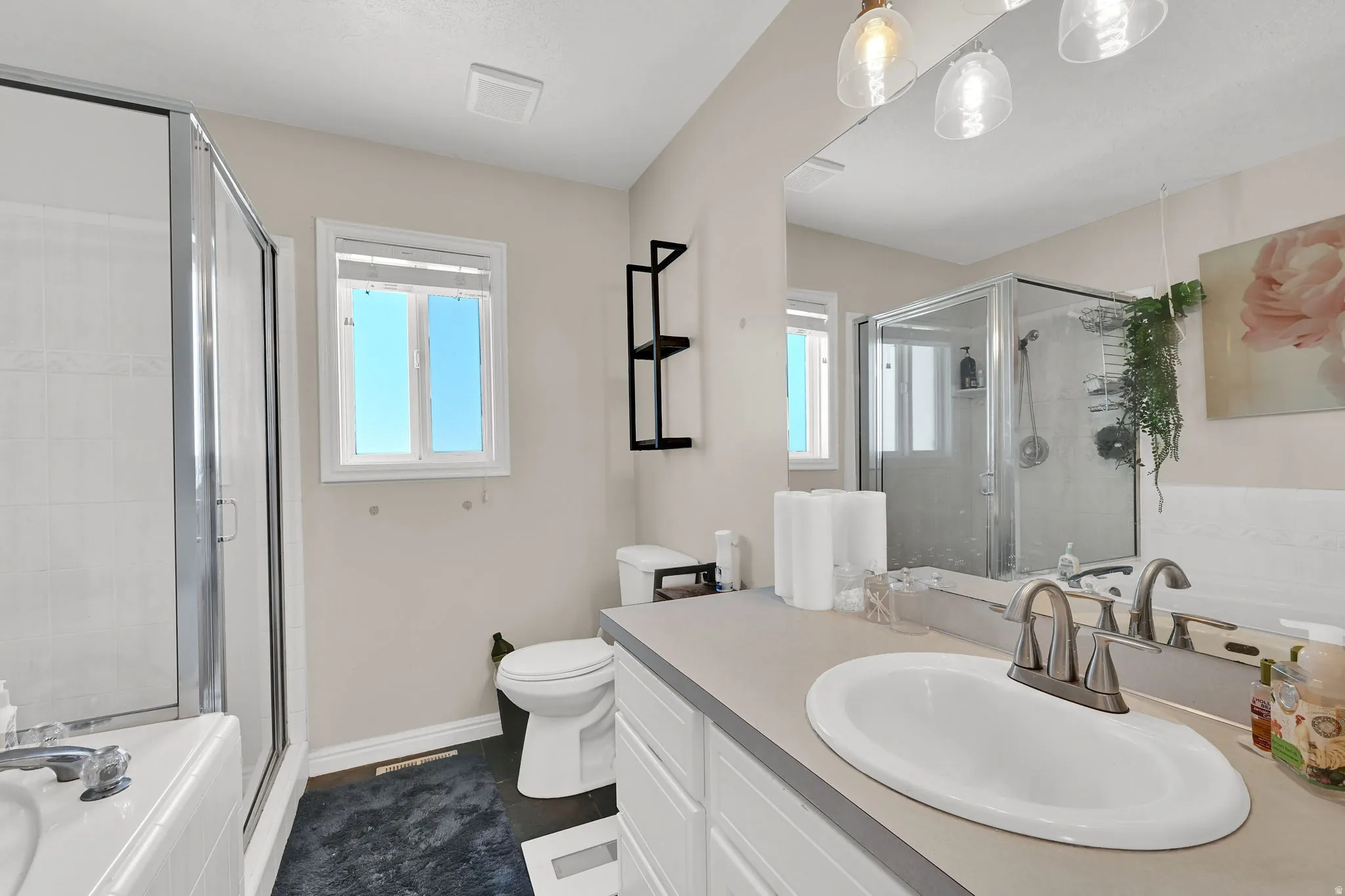 Full bathroom featuring a shower stall with clear glass enclosure, a built-in bathtub, a window with horizontal blinds, a vanity with an integrated sink and light gray countertop, and a wall-mounted mirror with clear glass pendant lighting