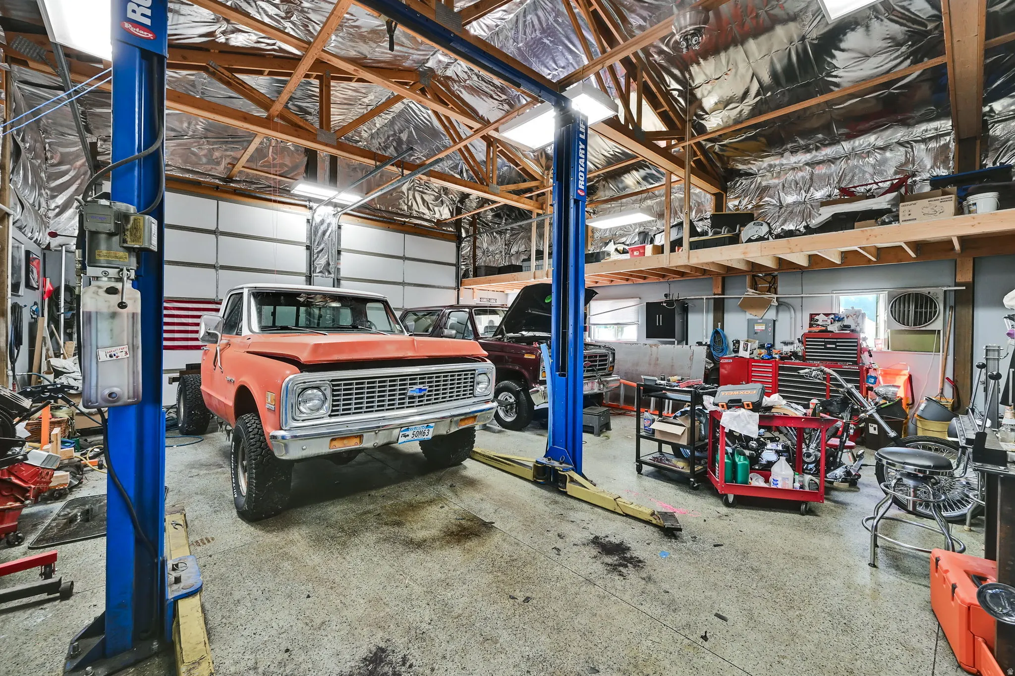 Spacious garage featuring a vehicle lift, exposed wooden truss ceiling, mezzanine storage, roll-up garage doors, and ample lighting