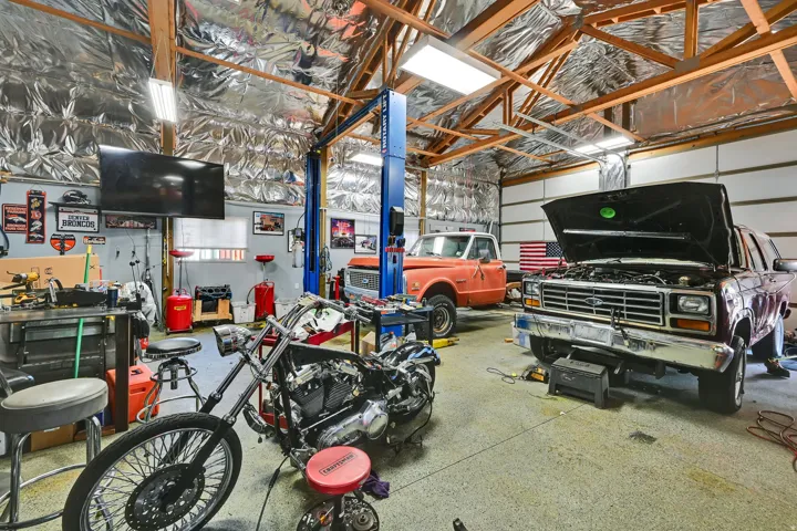 Spacious workshop with a vehicle lift, multiple garage doors, exposed wood beam ceiling, and overhead fluorescent lighting