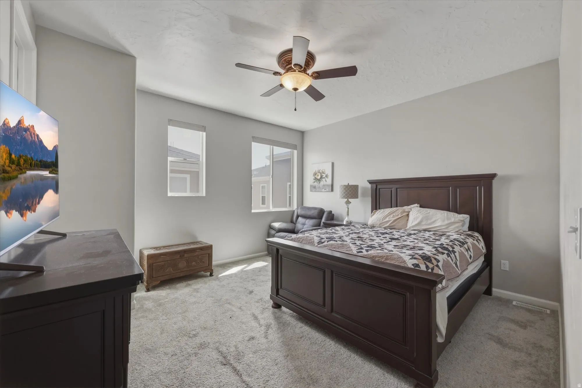 Carpeted bedroom featuring neutral wall tones, two windows with roller shades, and a ceiling fan with integrated lighting