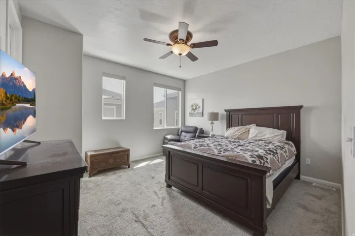 Carpeted bedroom featuring neutral wall tones, two windows with roller shades, and a ceiling fan with integrated lighting