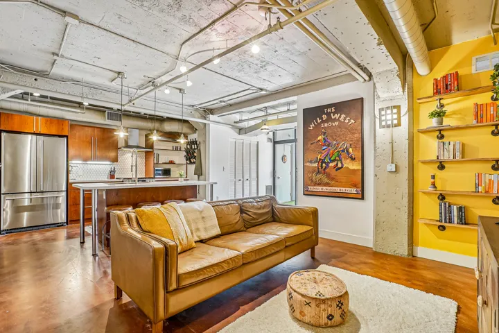 Open-concept living space featuring wood-finish flooring, exposed concrete ceiling with industrial lighting, and a kitchen with stainless steel appliances, wood cabinetry, and a white subway tile backsplash