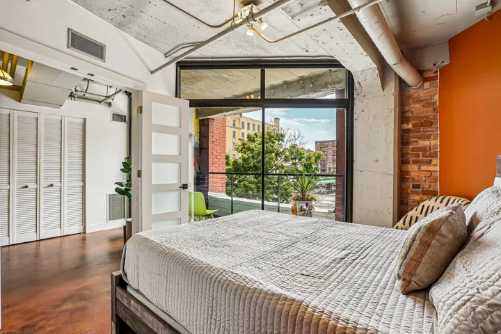 Expansive window wall with black framing, exposed brick accent wall, concrete ceiling with exposed piping, solid core door with frosted glass panels, and wood-finish flooring