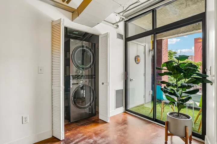 In-unit stacked laundry appliances within louvered bifold doors, concrete flooring, and a sliding glass door opening to a private patio