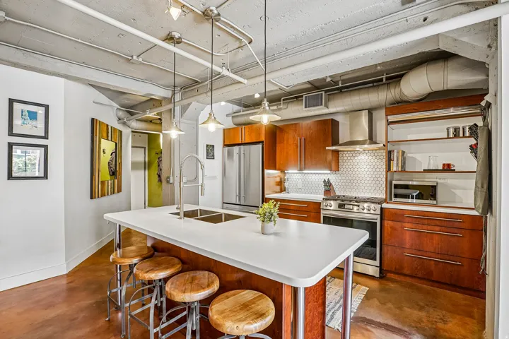 Modern kitchen featuring a large center island with integrated double sink, stainless steel appliances, wood cabinetry, hexagonal tile backsplash, and concrete flooring
