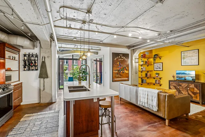 Open-concept living space featuring a kitchen island with dual sinks, warm wood-finish cabinetry, exposed industrial-style ceiling elements, and a wall of built-in shelving
