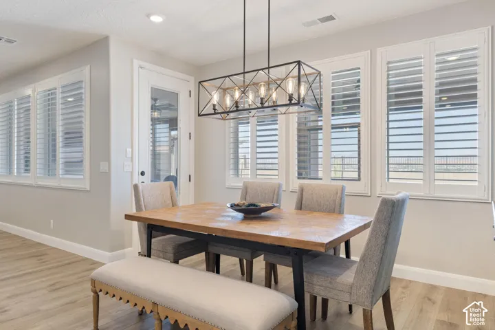Open-concept dining area featuring wood-finish flooring, a contemporary linear chandelier, multiple windows with white plantation shutters, a glass-paneled door, and neutral wall tones