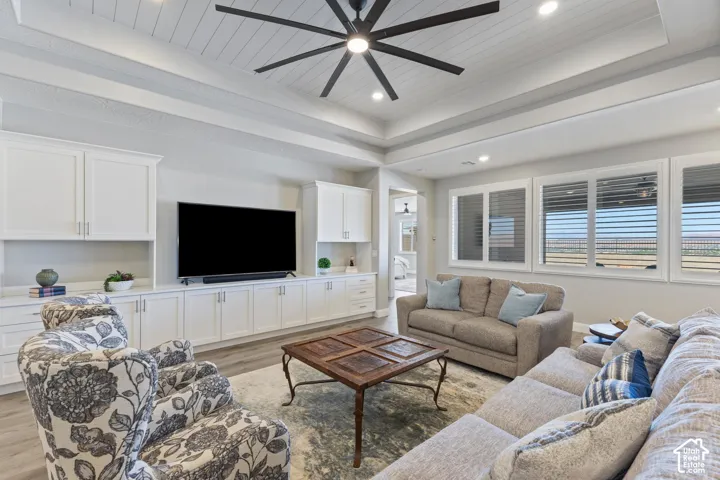 Living area featuring a coffered ceiling with a large ceiling fan, an extensive built-in media console with storage, multiple windows with blinds, and light wood-finish flooring