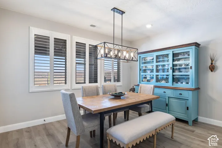 Dining area featuring engineered wood flooring, three windows with white plantation shutters, a linear chandelier with exposed bulbs, recessed ceiling lighting, and light gray walls