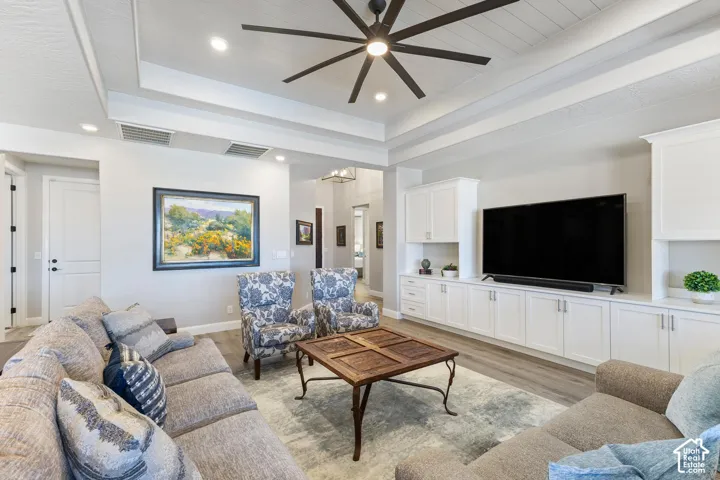 Living area featuring wood-finish flooring, a tray ceiling with a dark-bladed ceiling fan, recessed lighting, and built-in white cabinetry with a media center