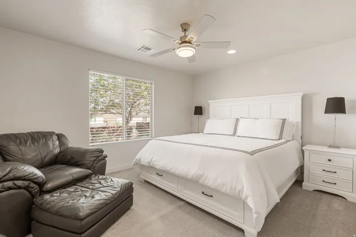 Carpeted bedroom featuring a window with blinds, white panel wainscoting style headboard, matching white nightstands with dark hardware, and a ceiling fan with integrated lighting