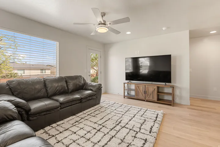 Living area featuring wood-finish flooring, a large window with horizontal blinds, a ceiling fan with integrated lighting, recessed lighting, and a glass-paneled door