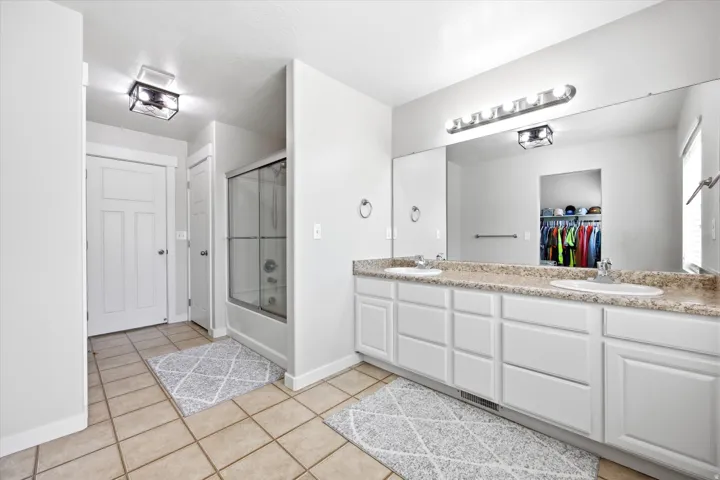 Bathroom featuring a dual vanity with a speckled countertop, a large wall-mounted mirror, white paneled cabinetry, and a shower with a sliding glass enclosure