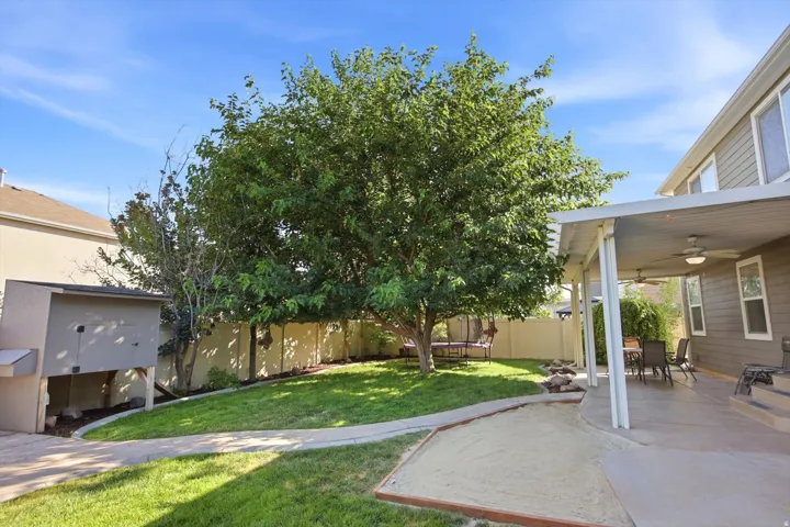 Expansive backyard featuring a mature central tree, covered patio with ceiling fans, concrete patio, sandpit, and manicured lawn