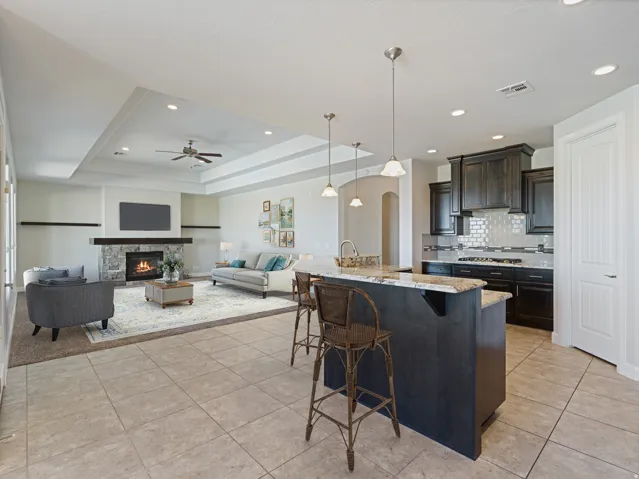 Open-concept living space featuring ceramic tile flooring, a stone fireplace with a wood mantle, and a tray ceiling with recessed lighting