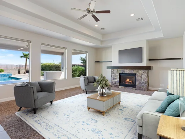 Living area featuring a stone-clad fireplace with wood mantle, multiple windows providing outdoor exposure, tray ceiling, recessed lighting, and a ceiling fan