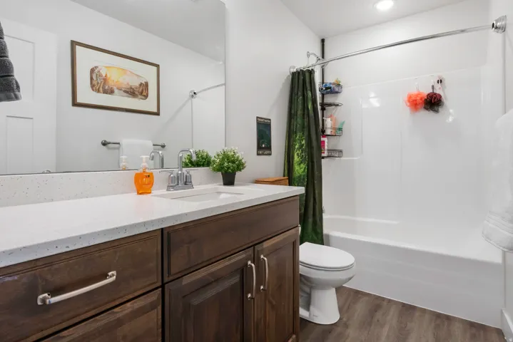 Wood-finish flooring, dark wood-tone vanity with chrome hardware, white countertop with integrated sink, chrome faucet, and a white tub with shower