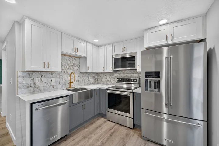 Kitchen featuring stainless steel appliances, two-tone cabinetry with gold-toned hardware, stone backsplash, white countertops, and wood-finish flooring